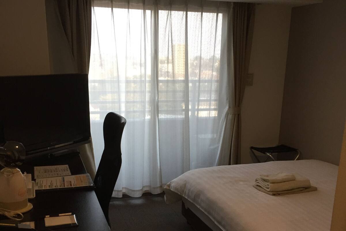 Een hotelkamer met een bed, een bureau met een computer, een stoel en een raam met gordijnen.