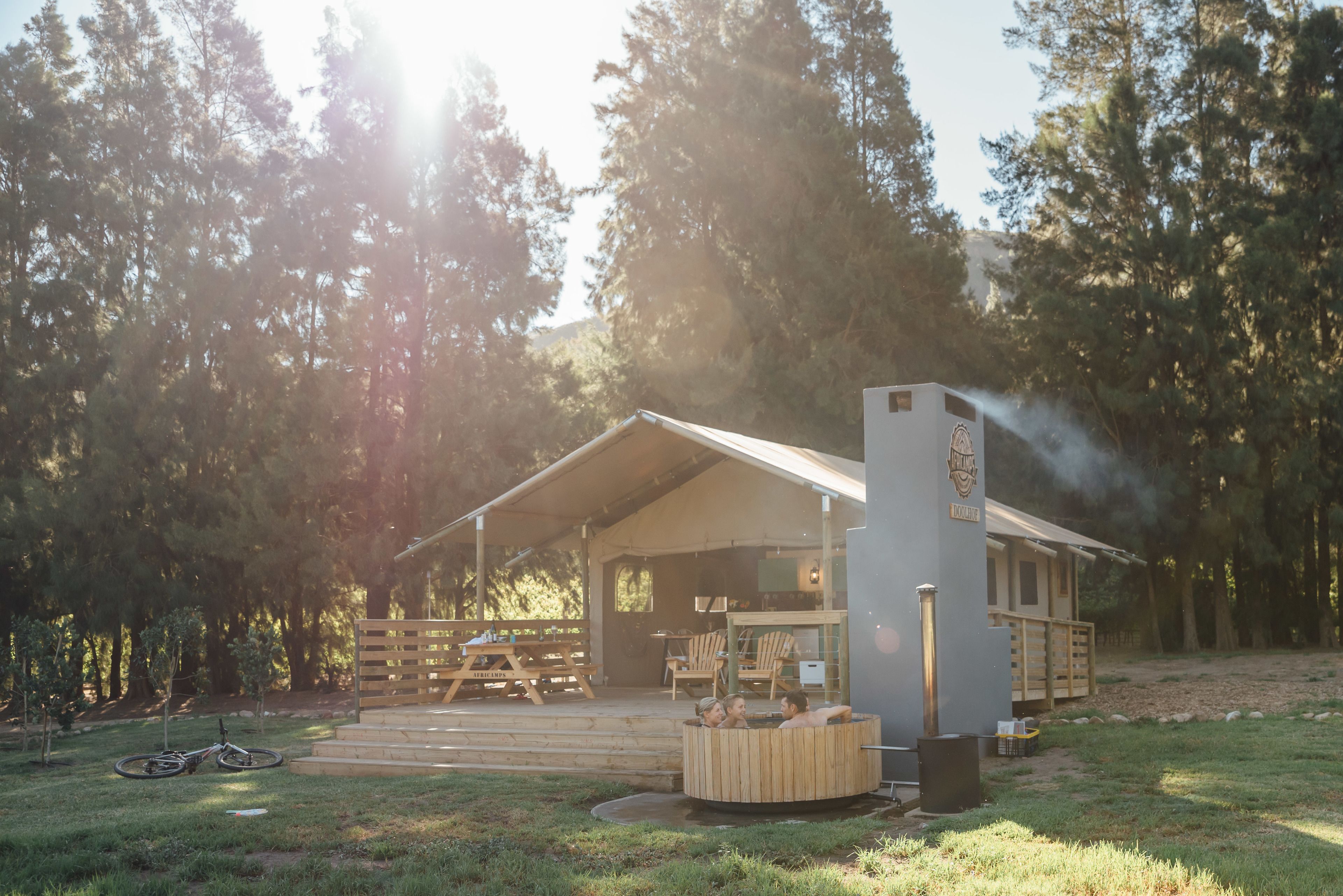 A rustic cabin with a hot tub, surrounded by trees, and a picnic area with benches and chairs.