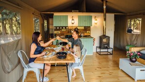 A family dining in a tented accommodation with a kitchen area, a wood stove, and a view of the forest.