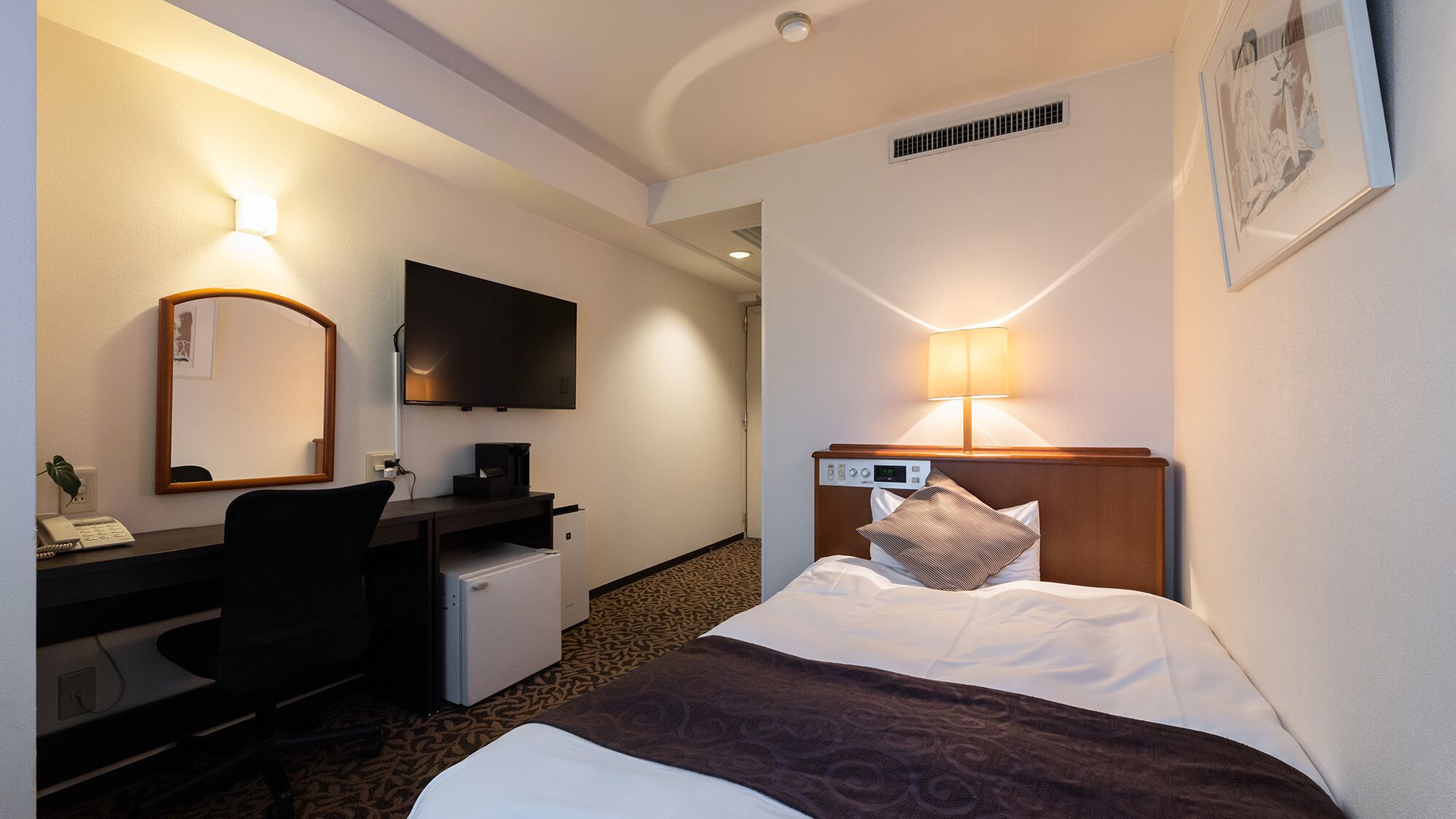 A hotel room with a bed, desk, chair, television, and a wall-mounted lamp.