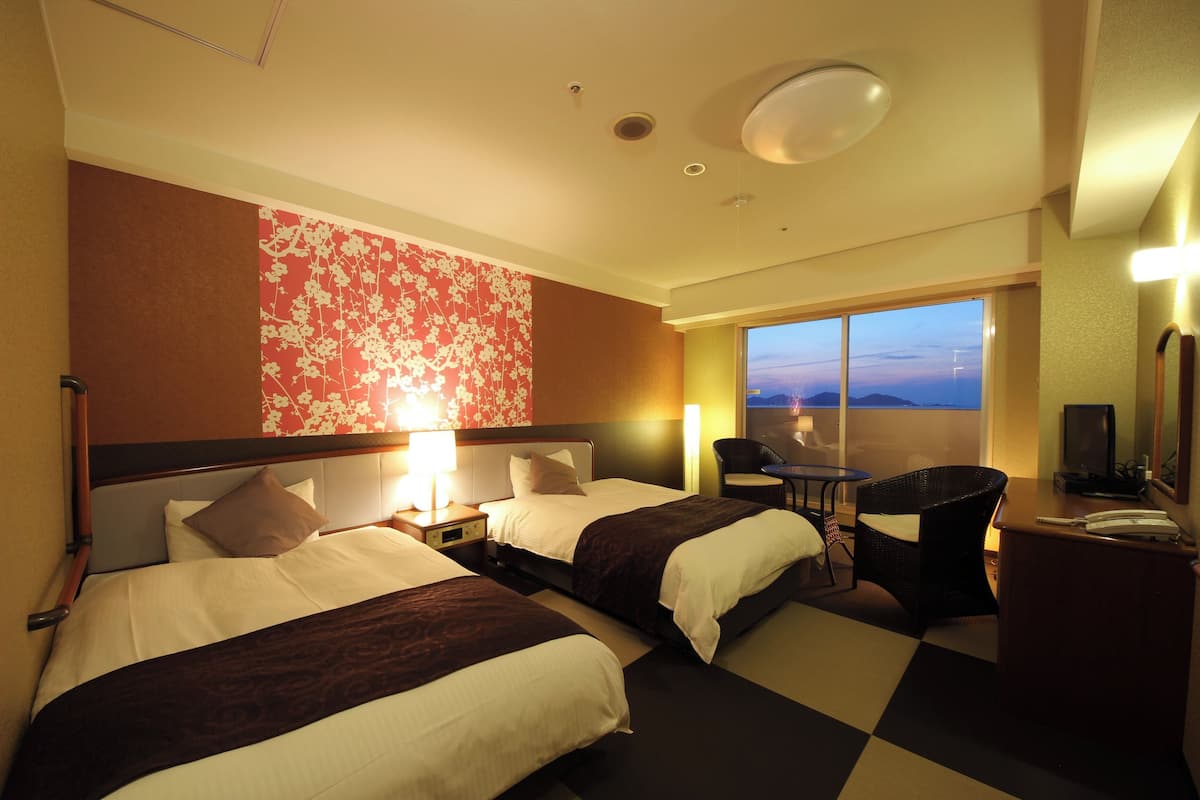 Habitación de hotel con dos camas, un escritorio, una silla, un televisor y un ventanal con vistas.