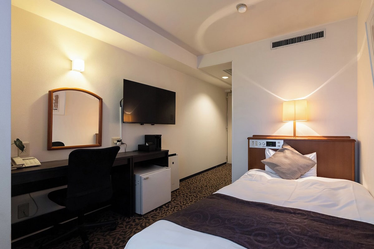 Una habitación de hotel con cama, escritorio, silla, televisor y un refrigerador pequeño.