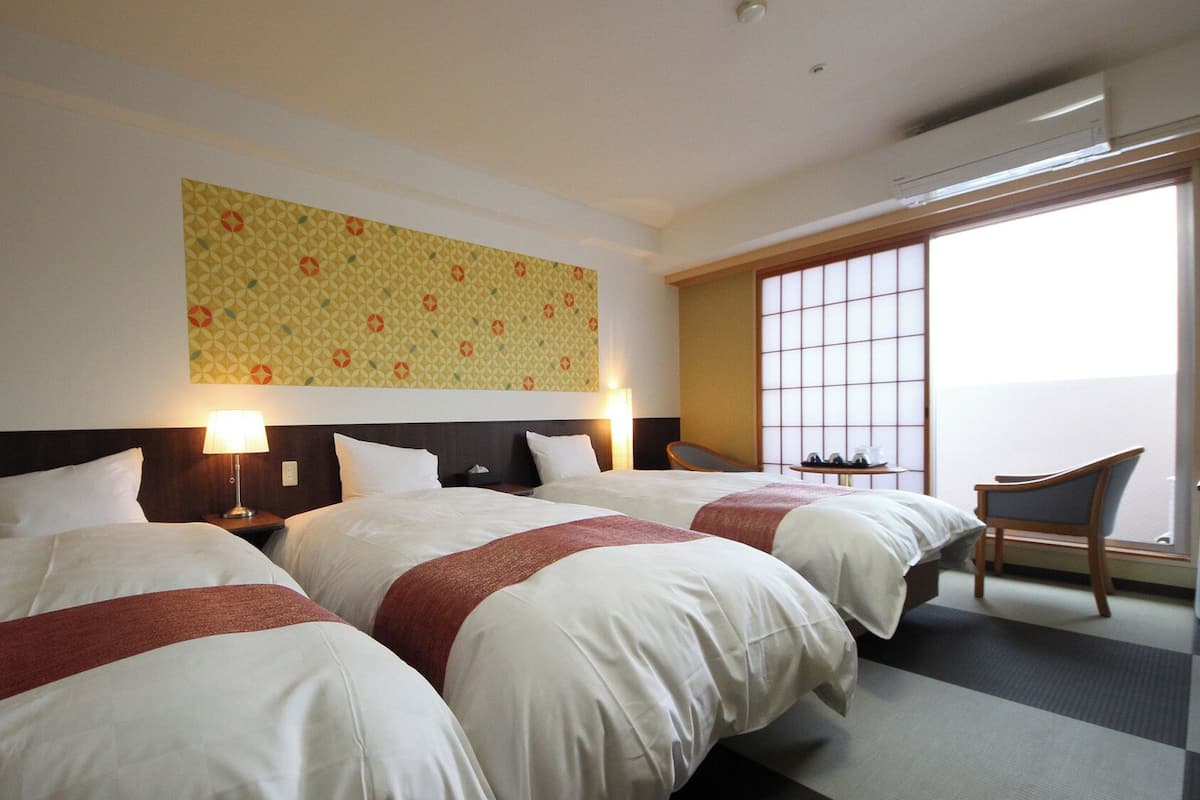 Habitación de hotel con dos camas, una silla, una mesa con lámpara y un ventanal grande.