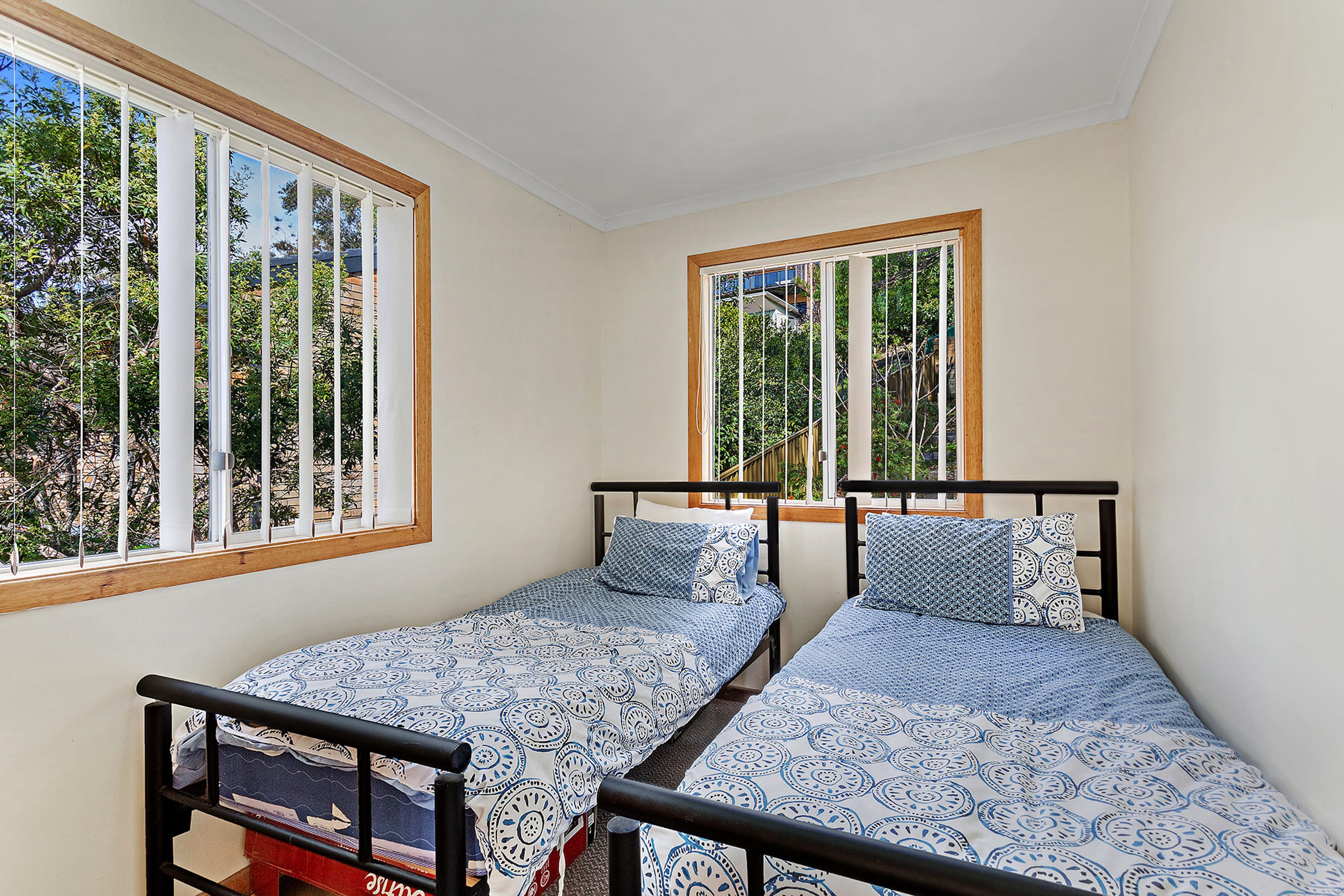 A room with two single beds, each with a patterned bedspread and pillows, positioned under windows with wooden frames.