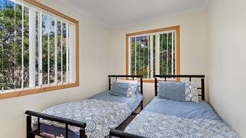 A room with two single beds, each with a patterned bedspread and pillows, positioned under windows with wooden frames.