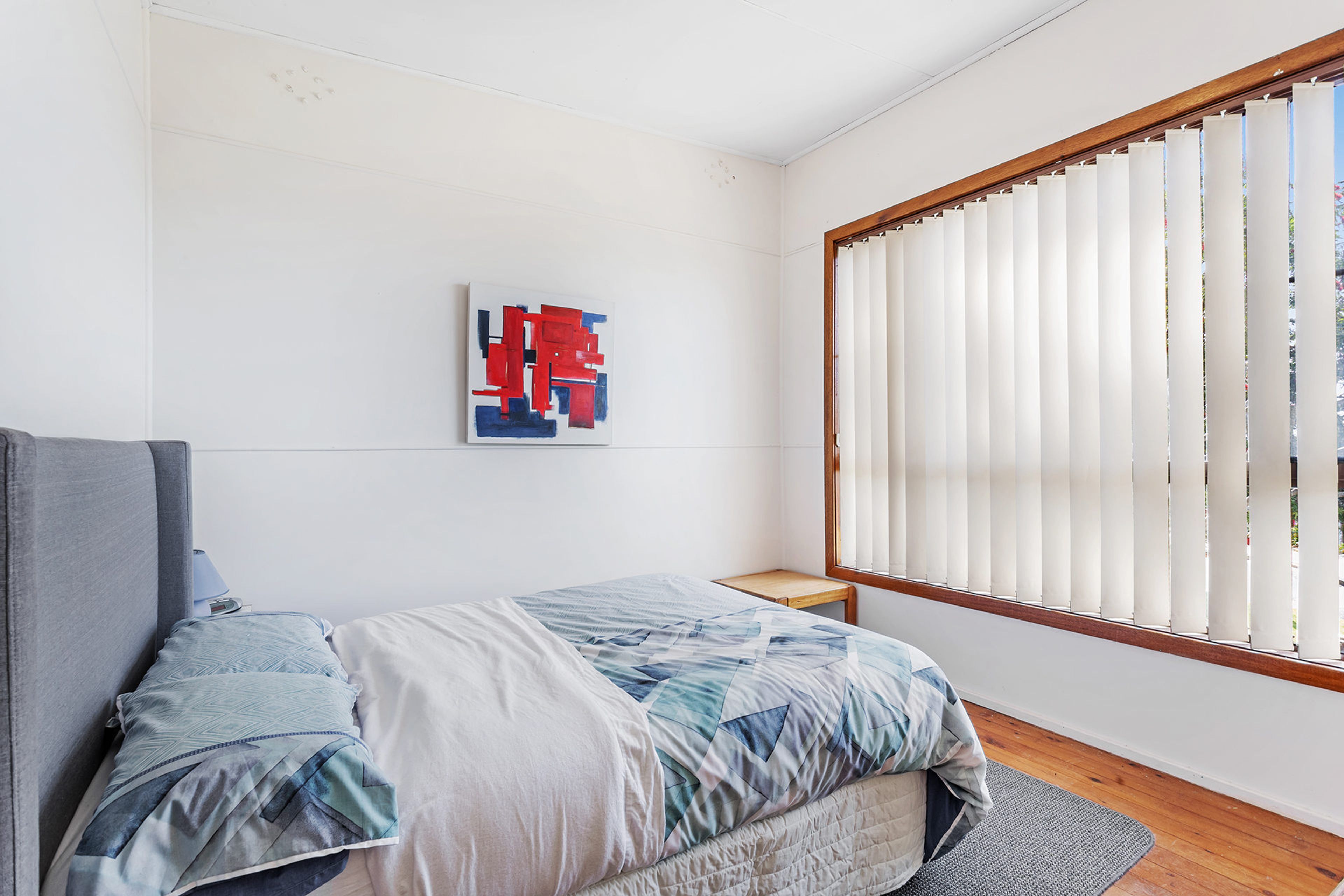 A bedroom with a bed, a window with blinds, a painting on the wall, and a small wooden stool.