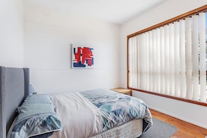 Een slaapkamer met een bed, een raam met luxaflex, een schilderij aan de muur en een klein houten krukje.
