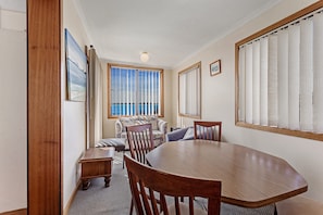 Een eethoek met een houten tafel en stoelen, een bank en uitzicht op de zee door grote ramen met zonwering.