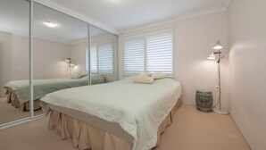 Un dormitorio con un espejo grande, una cama con un edredón de color claro, una mesita de noche con lámpara y una ventana con persianas.