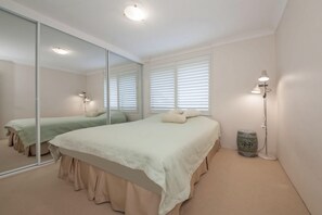 Een slaapkamer met een grote spiegel, een bed met een lichtkleurige dekbedovertrek, een nachtkastje met een lamp en een raam met jaloezieën.