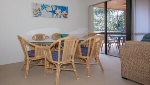 Un comedor con una mesa de vidrio y sillas de mimbre, un cuadro en la pared y vistas al exterior a través de un ventanal.