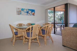 Een eetgedeelte met een tafel met glazen blad en rieten stoelen, een schilderij aan de muur en uitzicht op het landschap door een glazen deur.