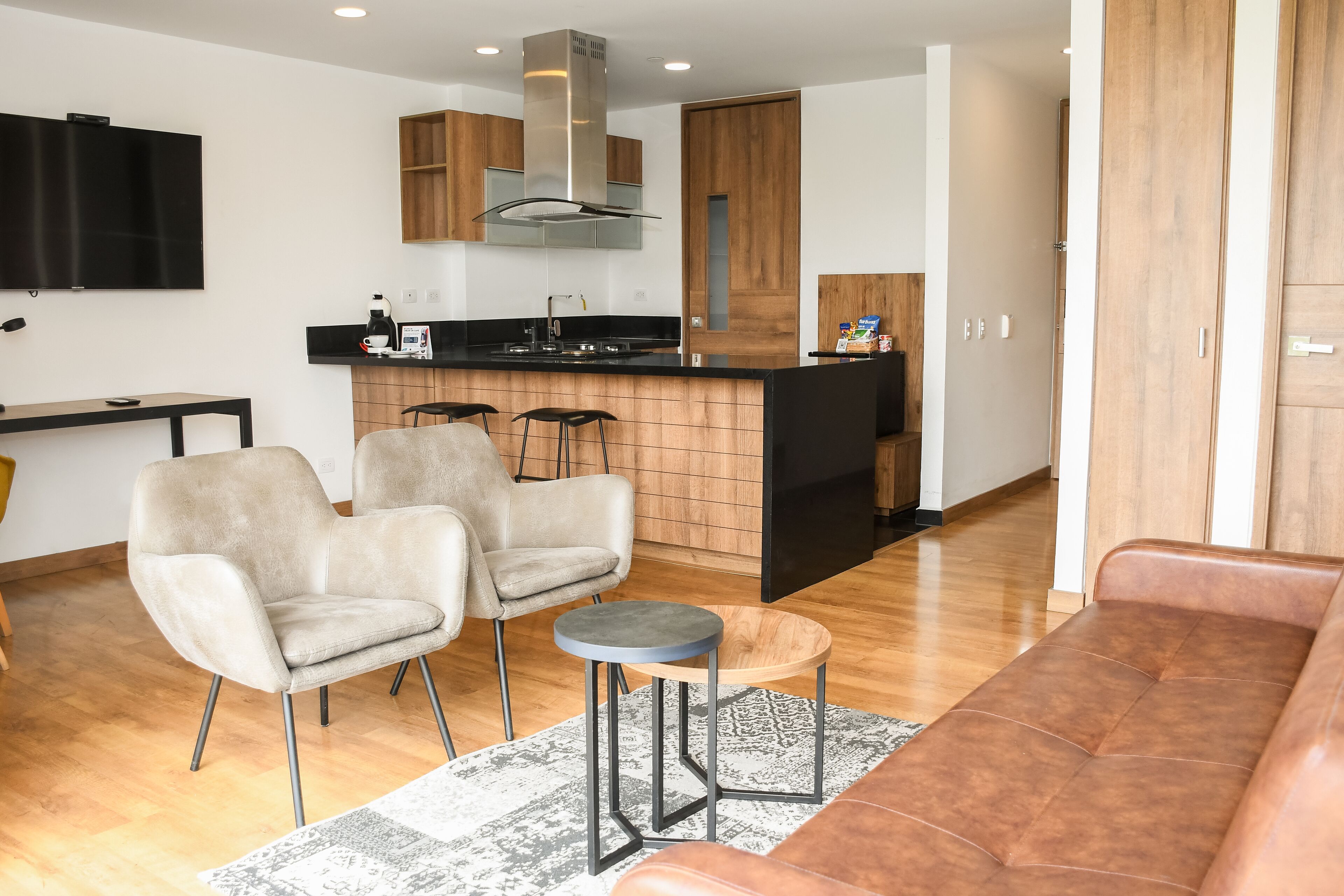 A modern kitchen with a central island, two bar stools, a sofa, and a TV mounted on the wall.