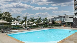A swimming pool with lounge chairs and umbrellas, surrounded by palm trees and a modern building. - Hotel Lagoon (Rionegro)