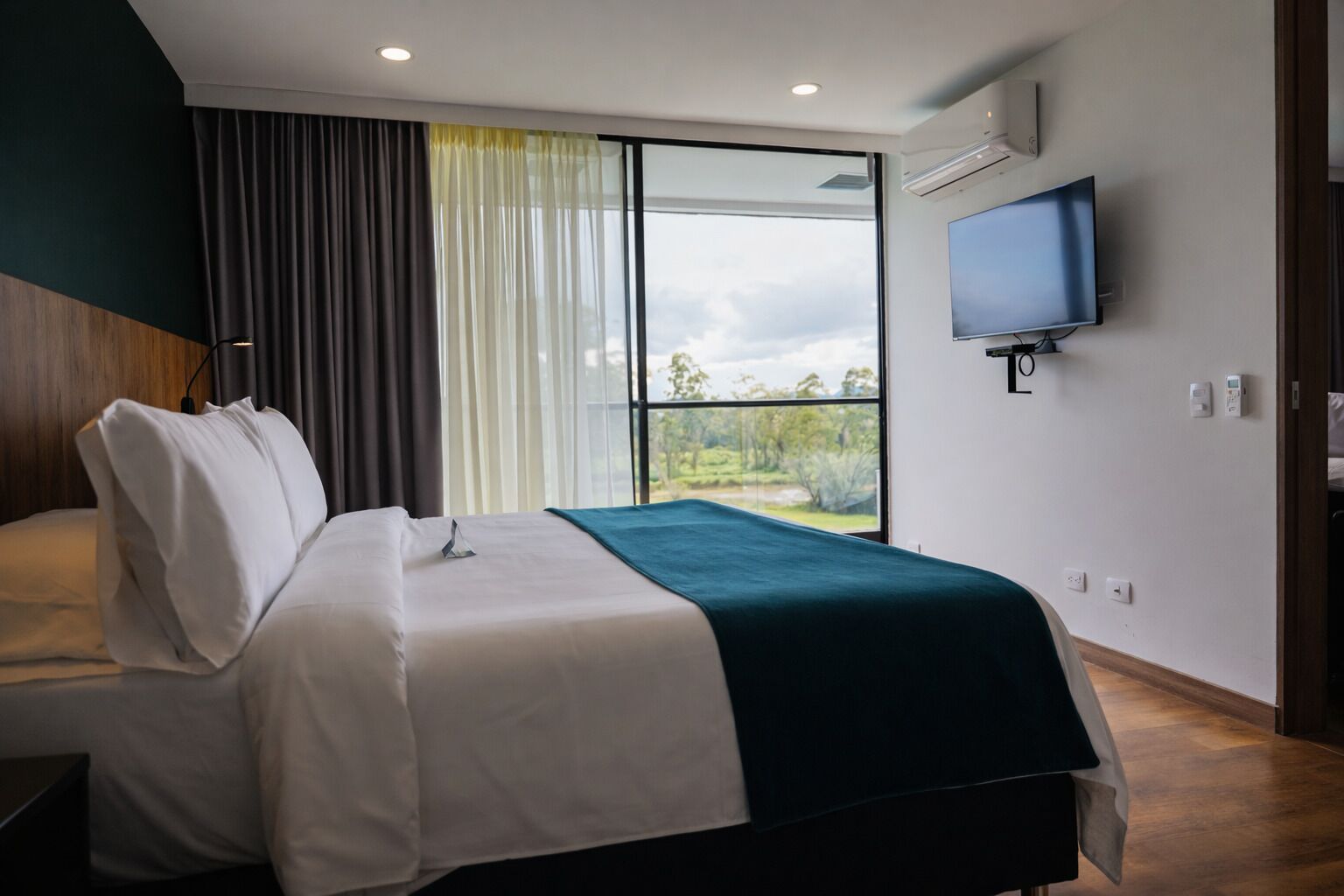 Una habitación de hotel moderna con una cama grande, un televisor montado en la pared y vistas al exterior.