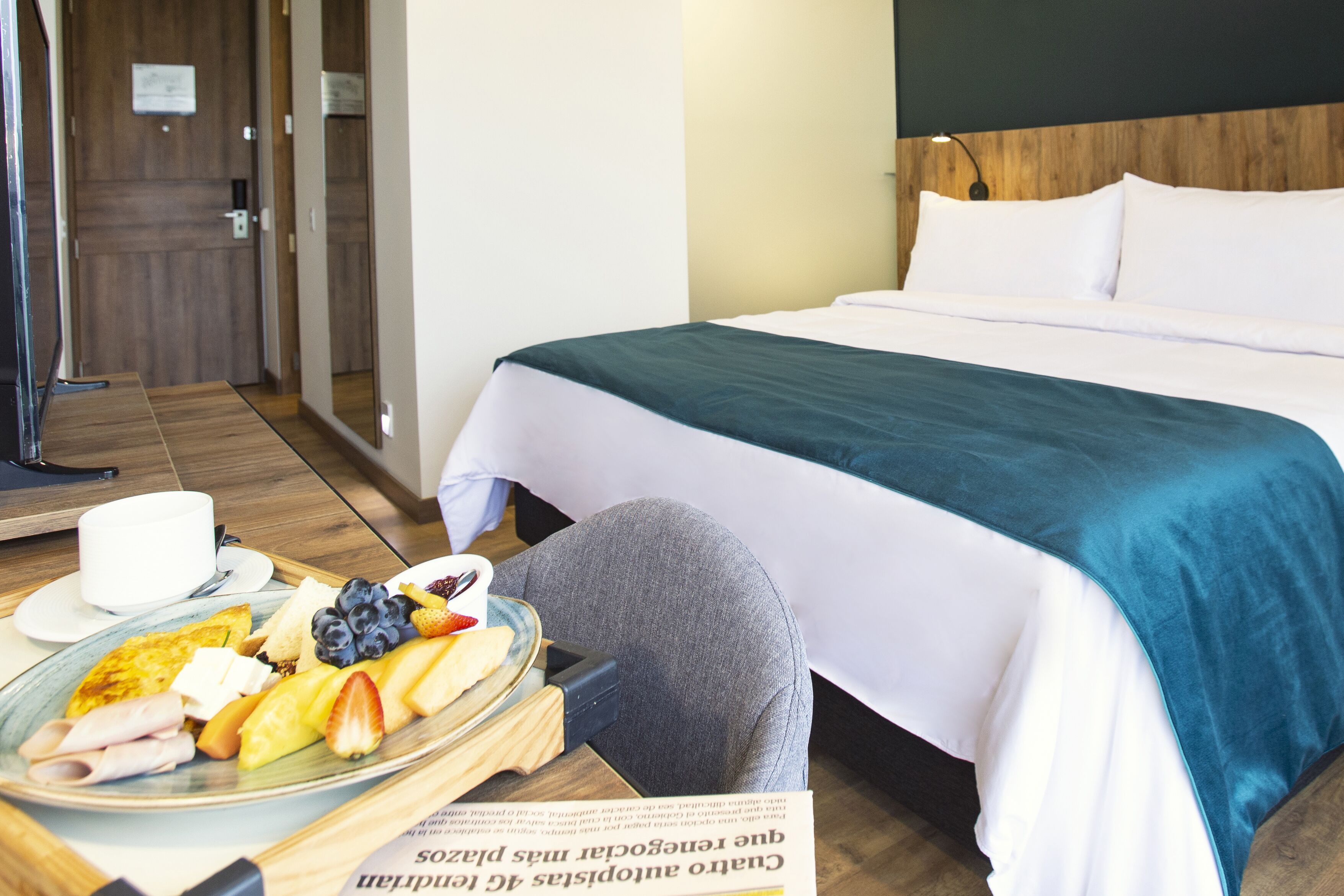 Ein Hotelzimmer mit einem Bett, einem Frühstücksbrett mit Früchten und Eiern sowie einer Zeitung.