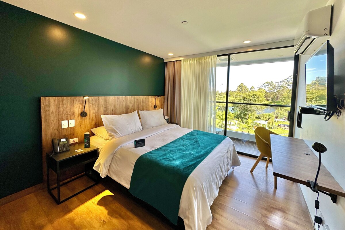 A modern hotel room with a large bed, a desk, a chair, and a view of the outdoors.