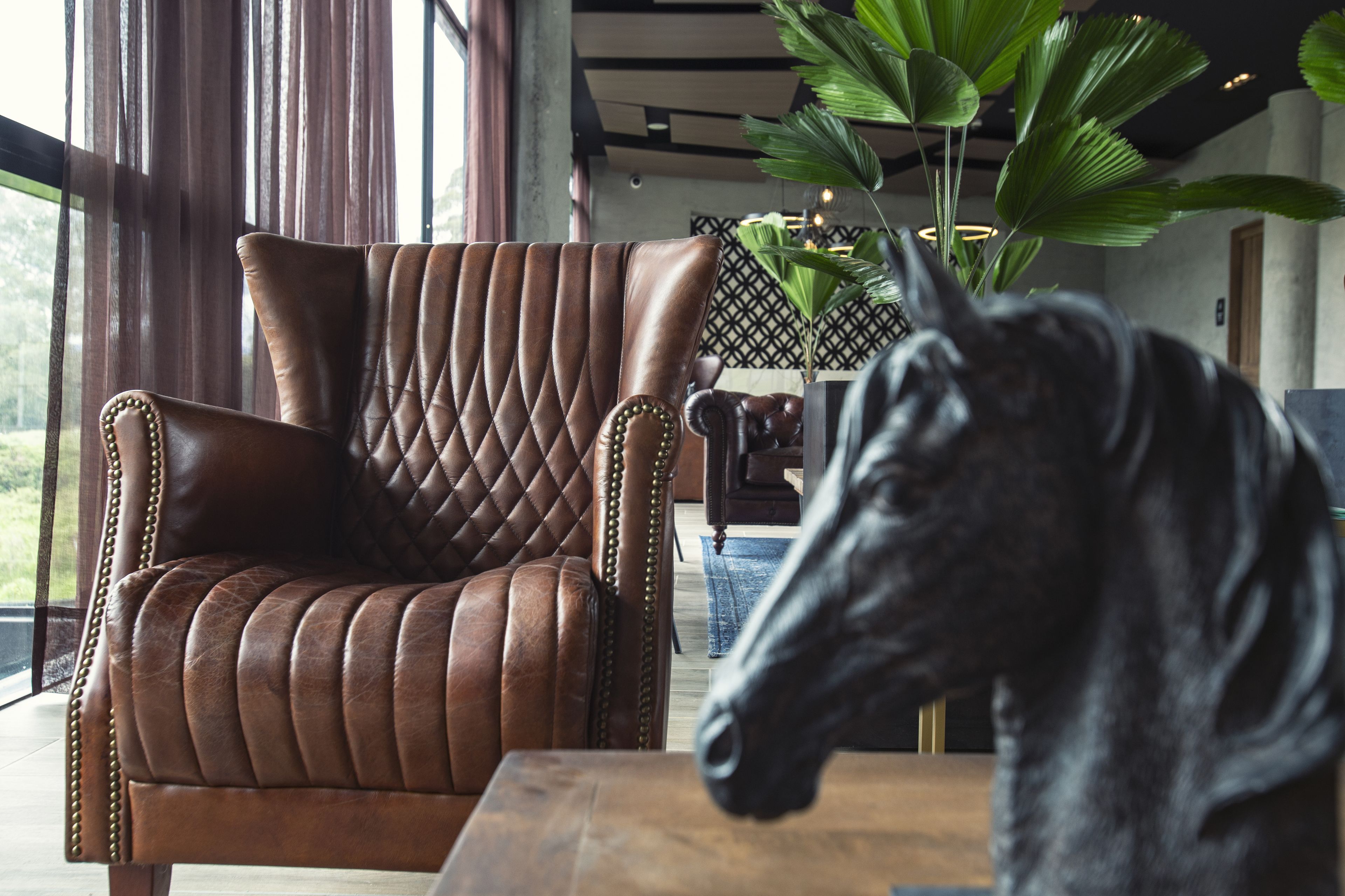 Un sillón de cuero marrón con respaldo acolchado y una mesa de madera en un interior moderno con ventanas amplias y una planta en maceta.