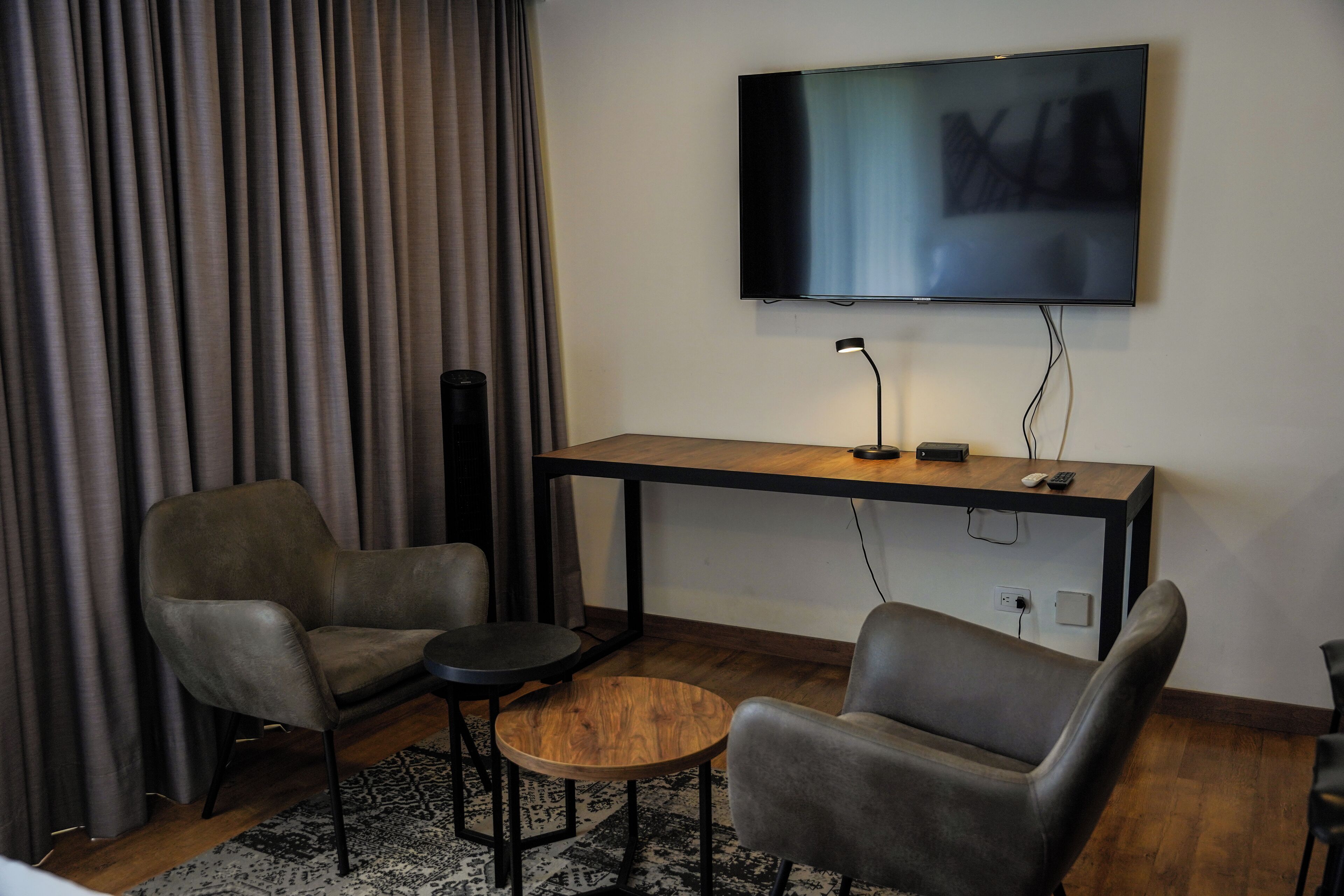 A modern living room with a flat-screen TV mounted on the wall, a wooden coffee table, two grey armchairs, and a dark floor lamp.