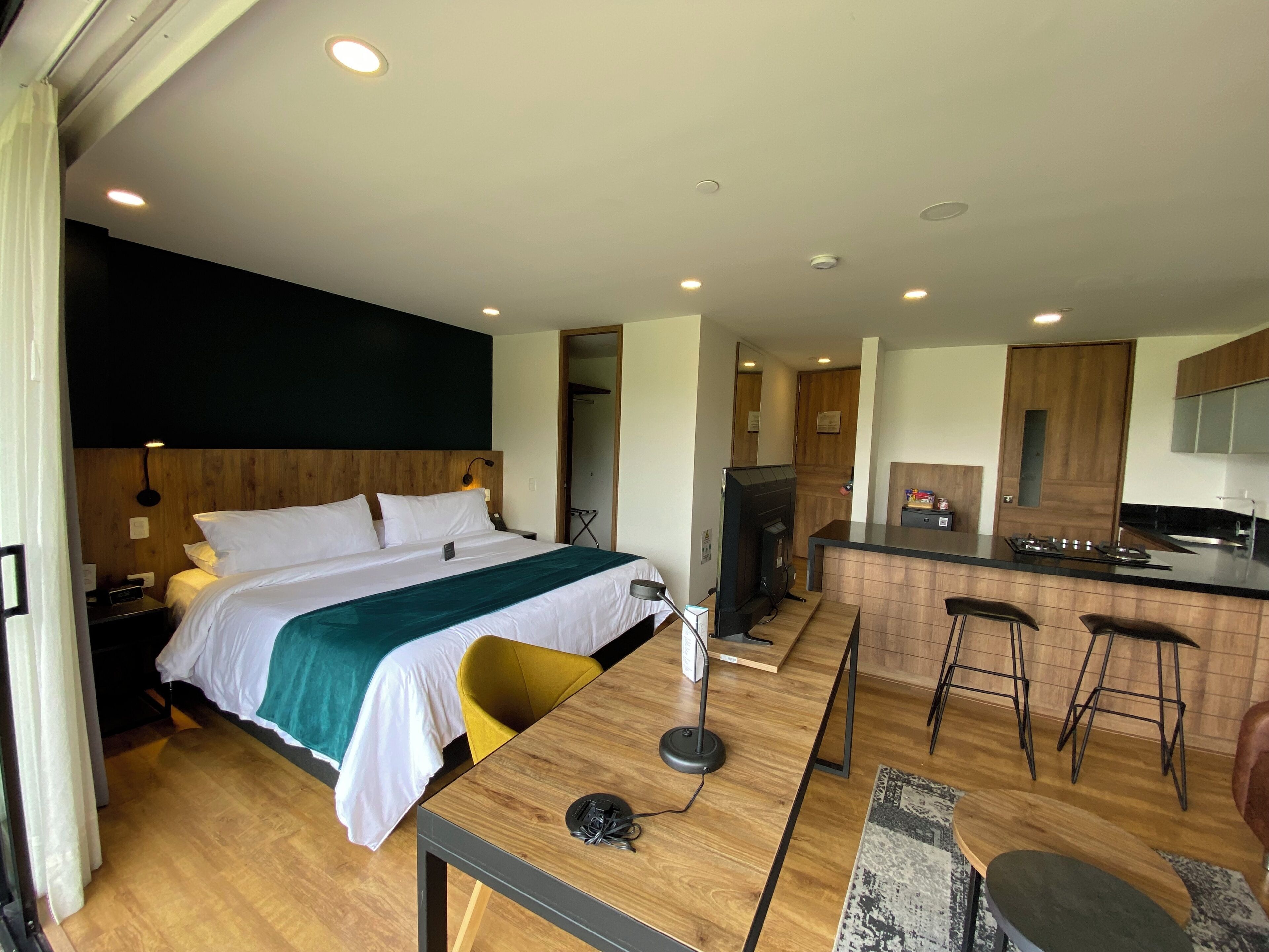 Una habitación de hotel moderna con una cama grande, un escritorio con una silla, una zona de cocina con estufa y taburetes altos, y un televisor pequeño.