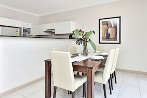A modern dining area with a wooden table set for four, white chairs, a vase with green plants, and a framed artwork on the wall.