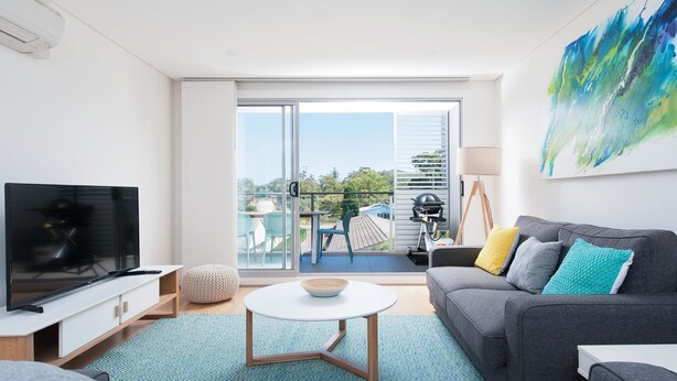 Ein modernes Wohnzimmer mit Flachbildfernseher, grauem Sofa mit bunten Kissen, rundem Couchtisch und einem Balkon mit Gartenmöbeln.