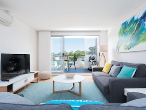 Een moderne woonkamer met een flatscreen-tv, een grijze bank met kussens, een ronde salontafel en uitzicht op het groen door een schuifdeur.