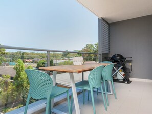 A balcony with a wooden table and four green chairs, a barbecue grill, and a view of a residential area.