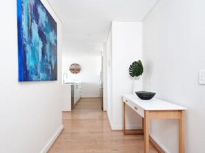 Un pasillo con piso de madera, pared blanca, un cuadro, un reloj, un lavamanos, un armario, una planta y una mesa.