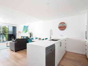 Eine moderne Küche mit Geschirrspüler, eine Essecke mit Tisch und Stühlen, ein graues Sofa und ein großes Fenster.