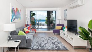 En moderne stue med en grå sofa, et fladskærms-tv og en balkon med havudsigt.