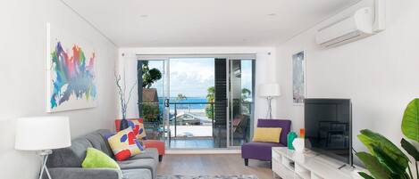 A modern living room with a grey sofa, a flat-screen TV, and a balcony with a view of the sea.