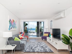 Een moderne woonkamer met een grijze bank, een flatscreen televisie en een balkon met uitzicht op zee.