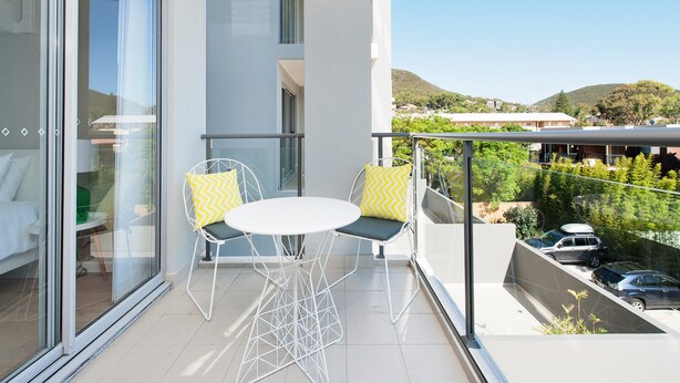 En balkon med en glasdør, der fører til et værelse, et lille bord med to stole og udsigt over en parkeringsplads og grønt område.