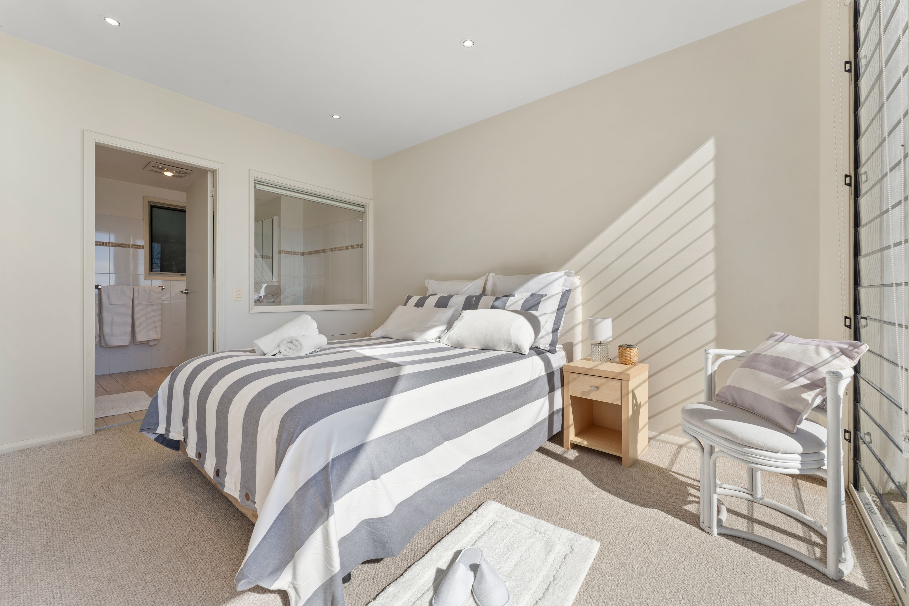 Une chambre à coucher comprenant un lit, une table de chevet, une chaise, un miroir et donnant sur une salle de bain.