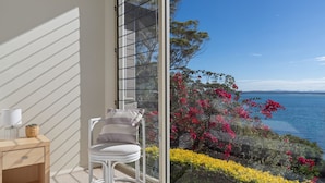 Un balcon avec une chaise, une petite table avec une lampe et une vue sur une étendue d’eau bordée de fleurs.