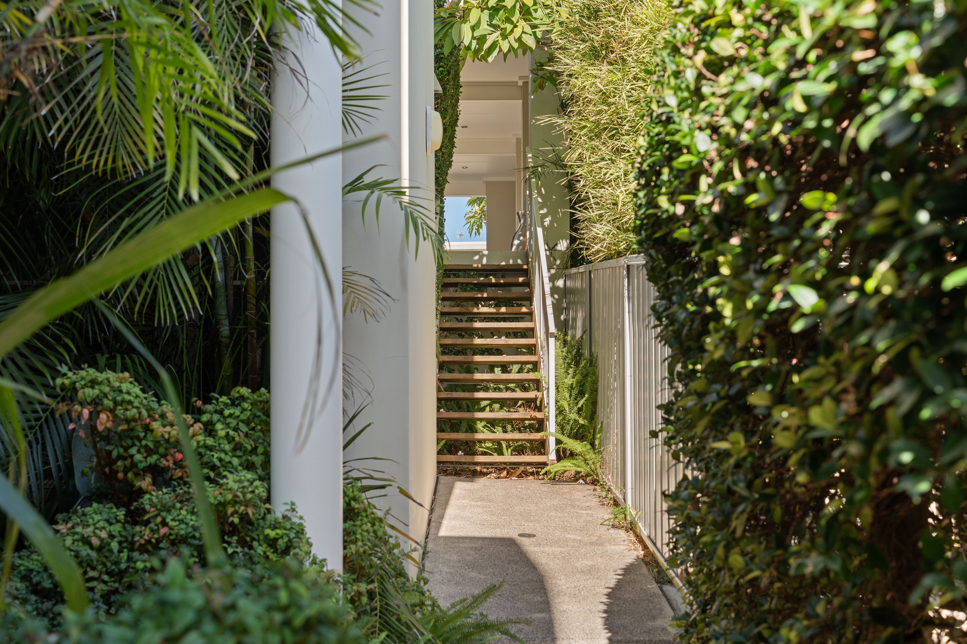Un étroit sentier mène à un escalier bordé d’une végétation luxuriante et à une structure de bâtiment blanche.