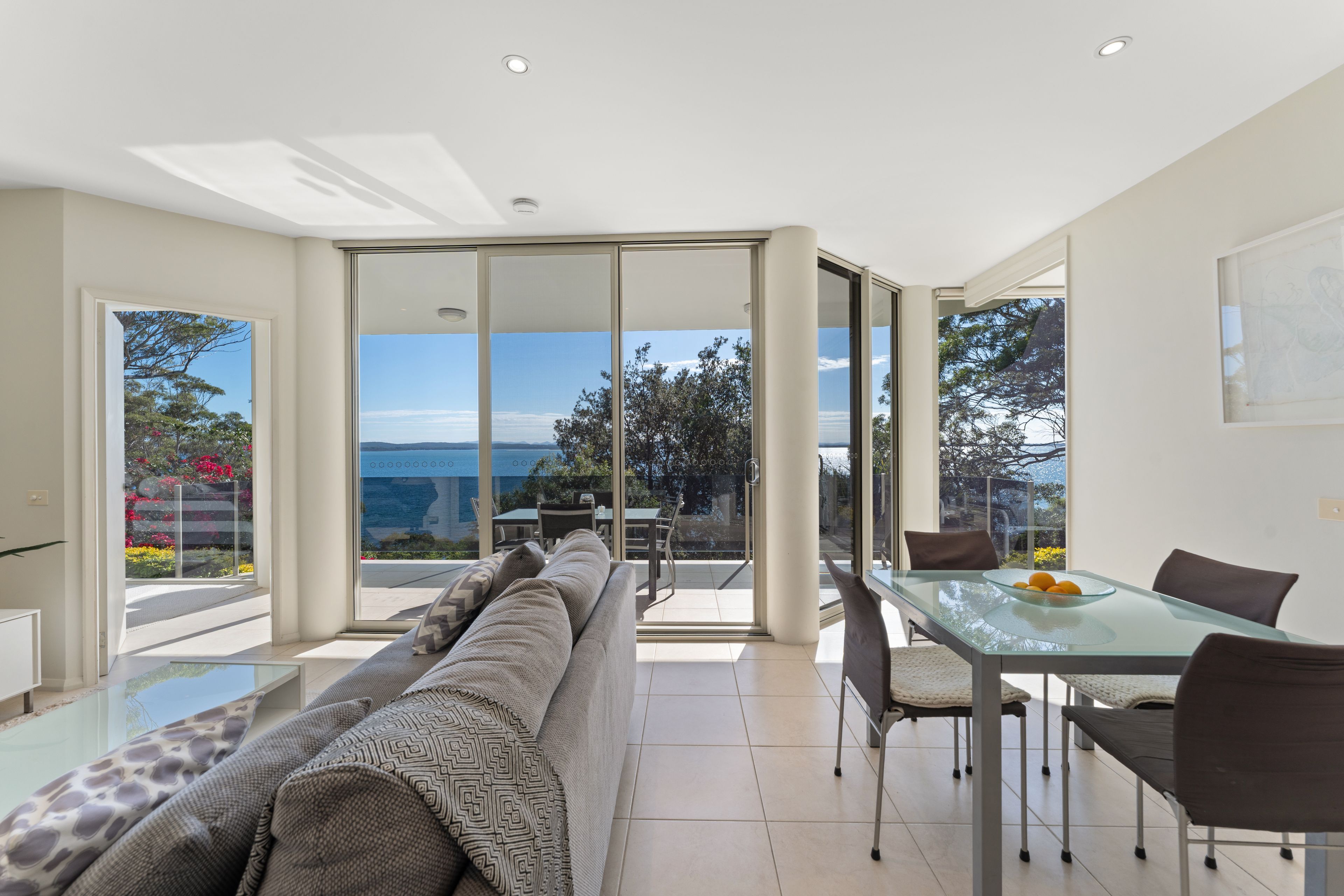 A modern living room with a glass dining table, a sofa, and a view of the outdoors.