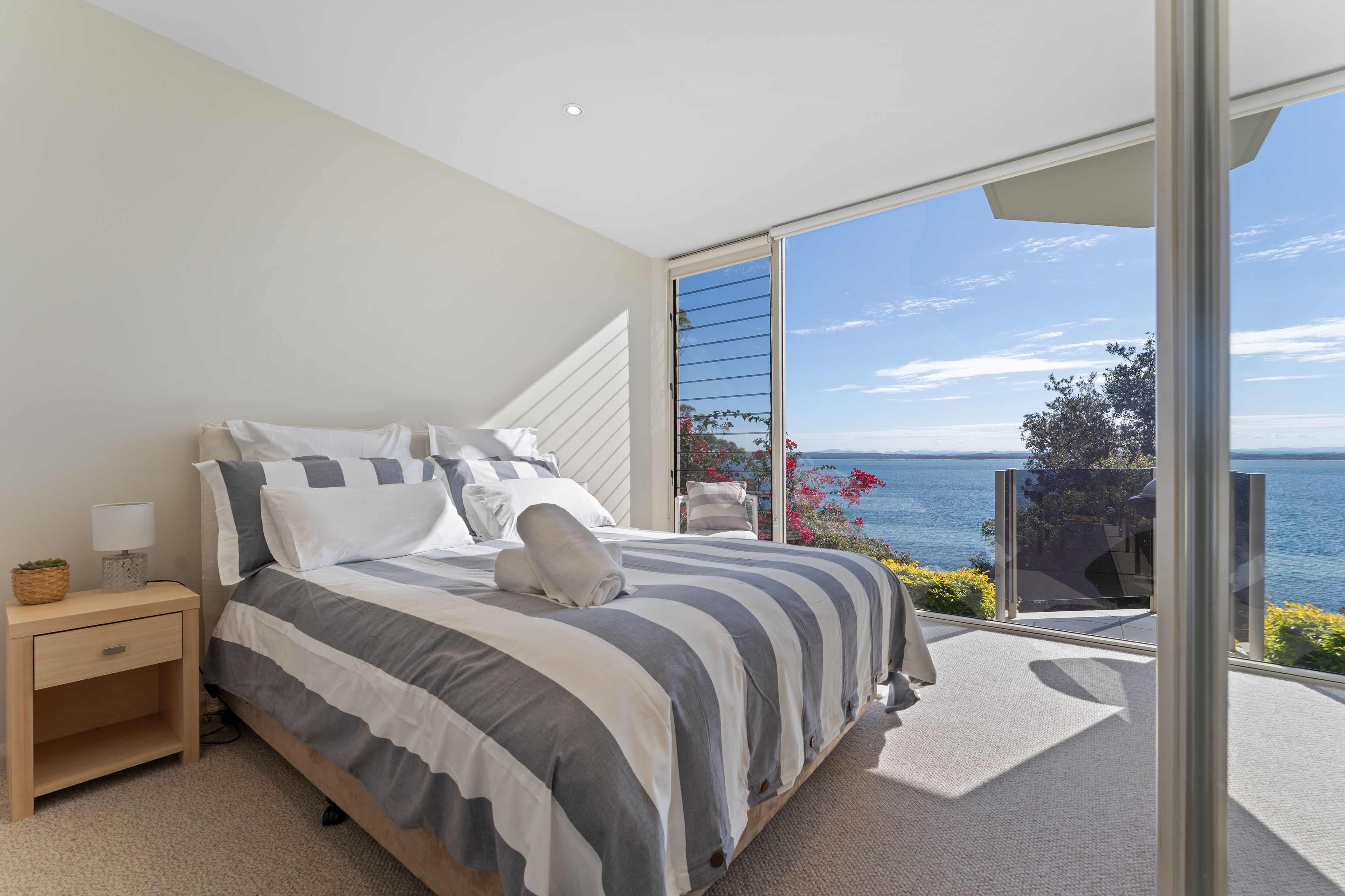 A bedroom with a large bed, a nightstand, and a view of the sea through a sliding glass door.