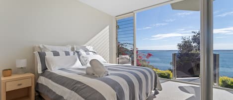 A bedroom with a large bed, a nightstand, and a view of the sea through a sliding glass door.