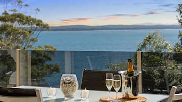 A table with a bottle of champagne, two glasses, and a decorative bowl with stones, overlooking a body of water with a clear sky.
