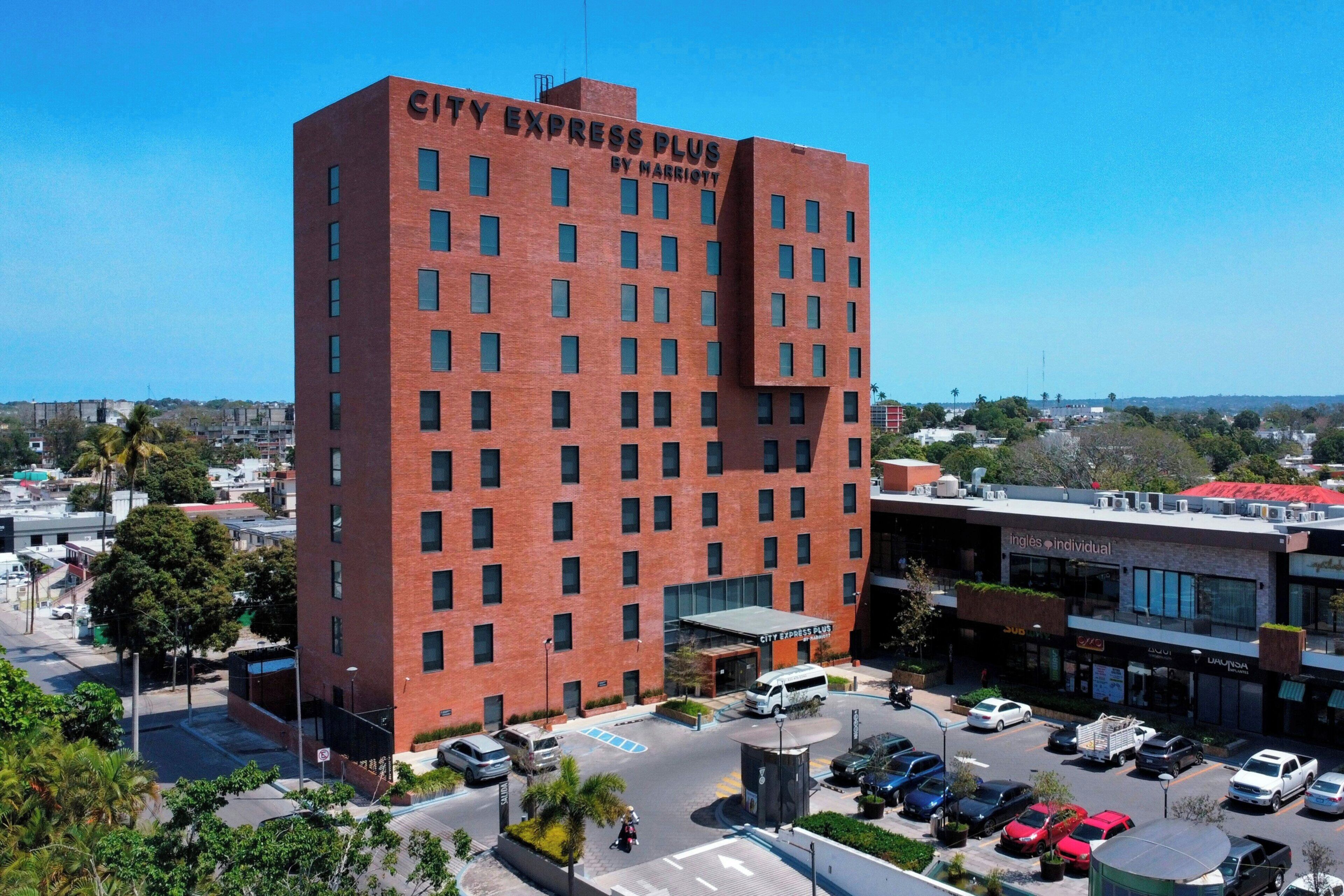 A multi-story hotel with a prominent City Express Plus sign, a parking lot, and a view of the surrounding cityscape.
