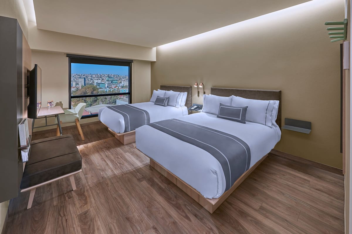 A hotel room with two beds, a desk, a chair, a TV, and a large window offering a city view.