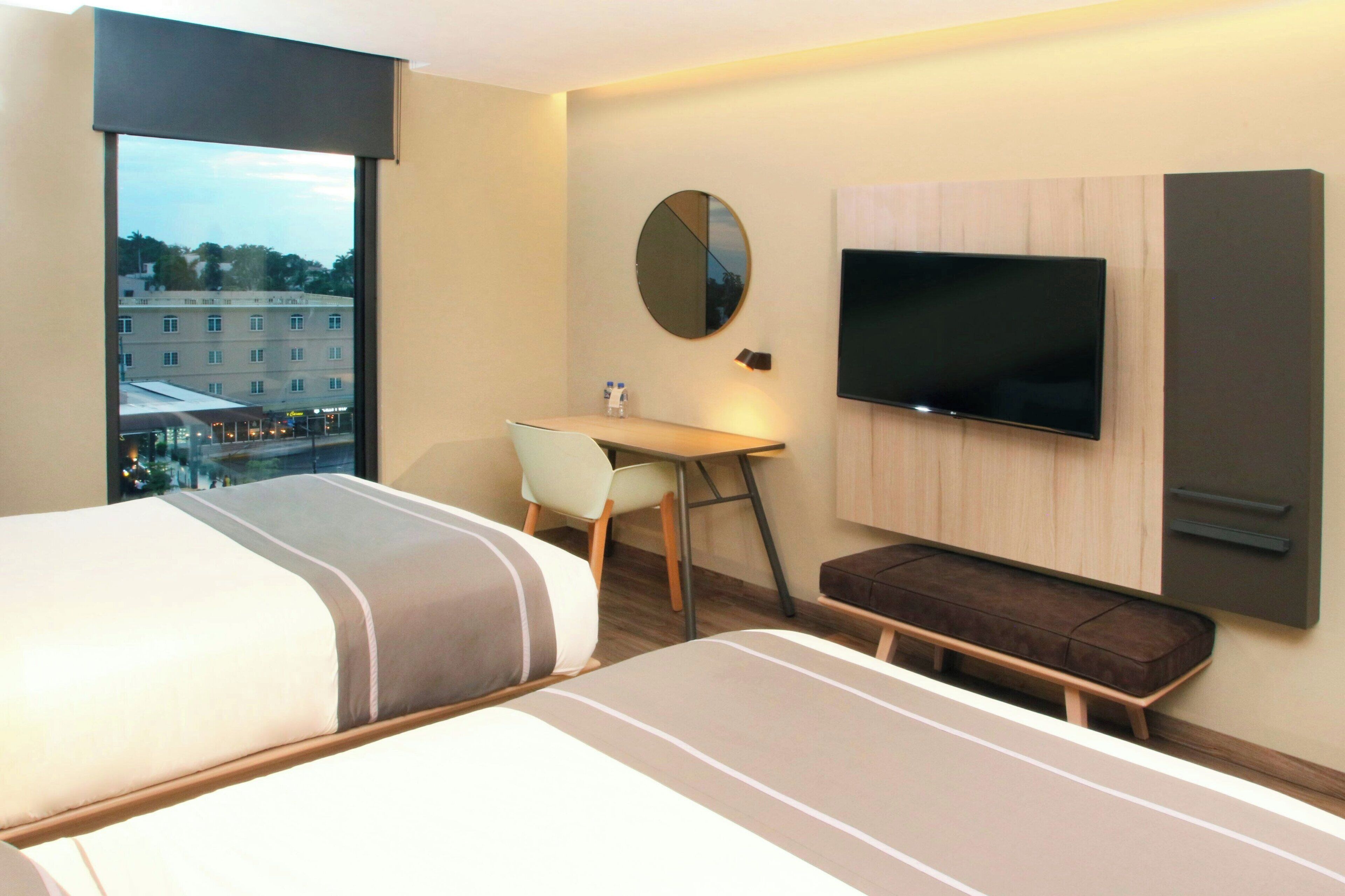Habitación de hotel con cama, escritorio, silla, televisor y vista a la ciudad.
