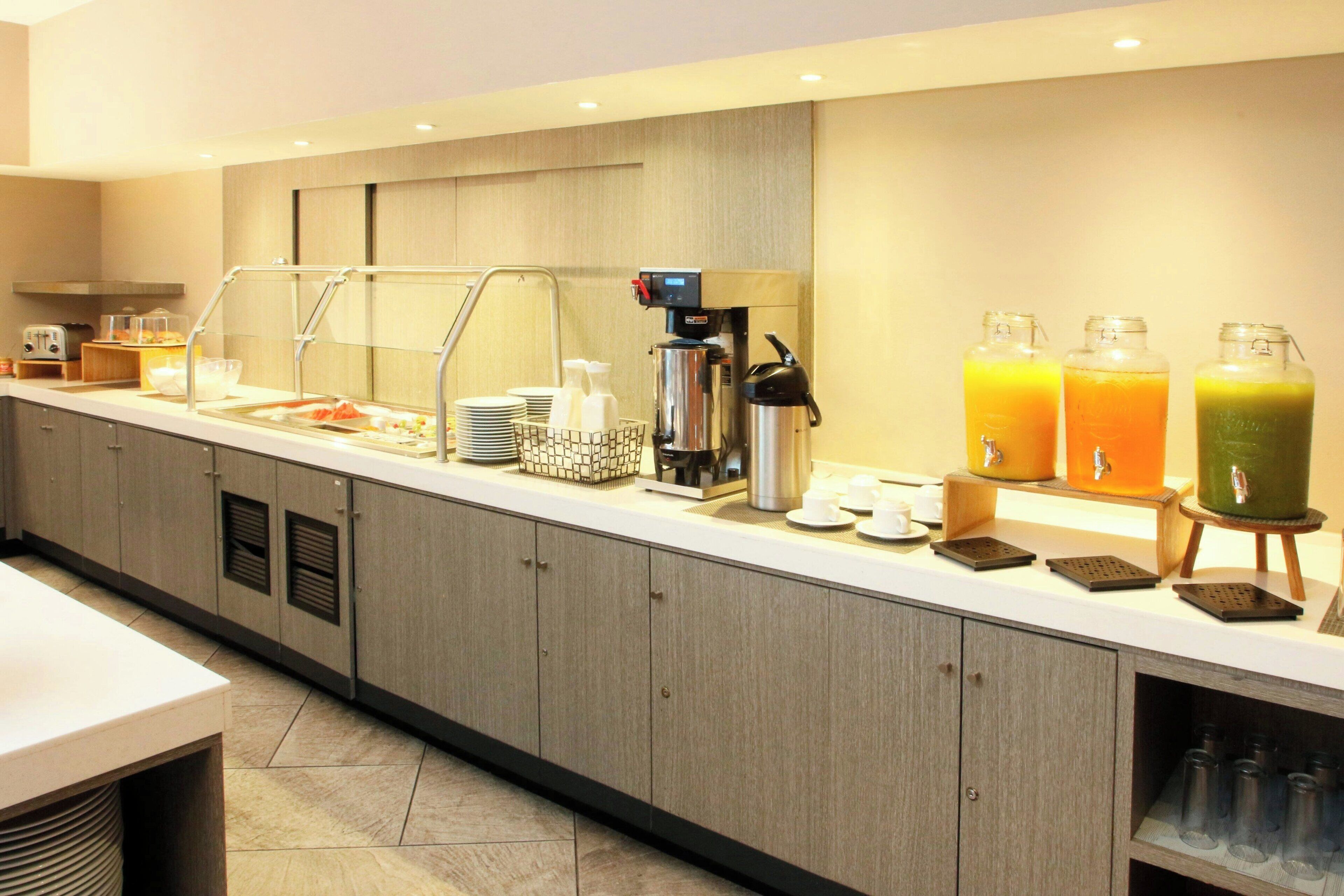 Un área de desayuno de hotel moderna con un mostrador de autoservicio, que incluye una máquina de café, dispensadores de jugo y una variedad de alimentos para el desayuno.