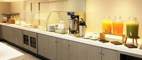 Un área de desayuno de hotel moderna con un mostrador de autoservicio, que incluye una máquina de café, dispensadores de jugo y una variedad de alimentos para el desayuno.