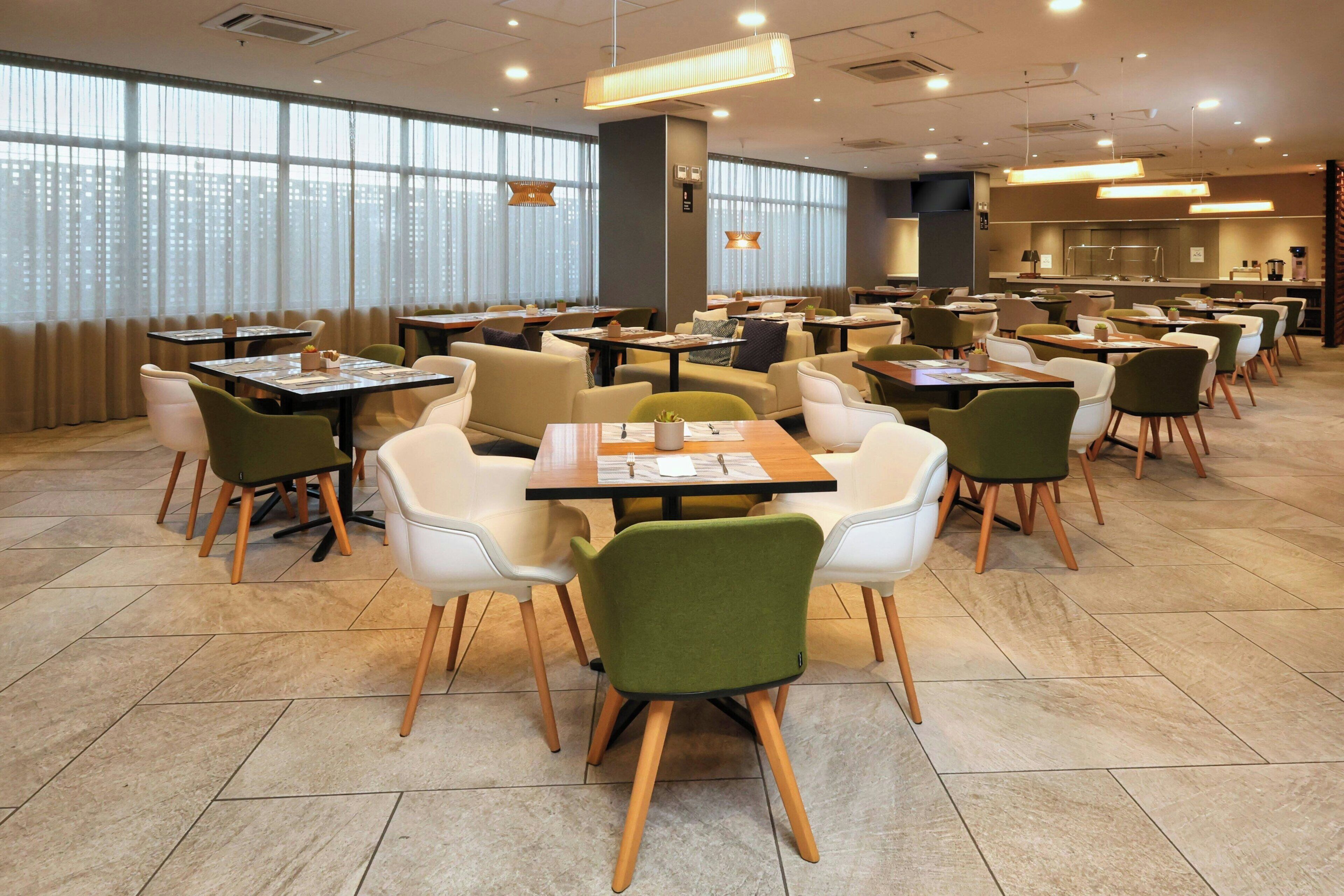 A spacious dining area with multiple tables and chairs, large windows with curtains, and a modern kitchen in the background.