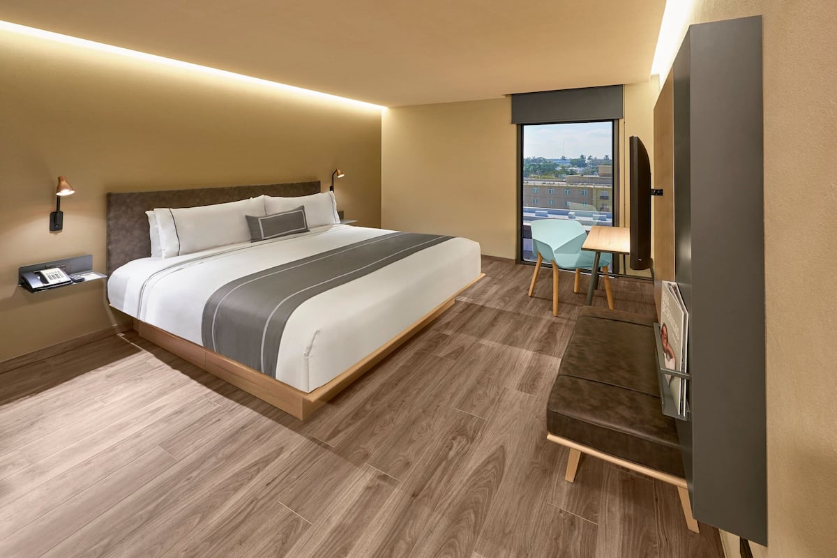 A modern hotel room with a large bed, a desk with a chair, and a large window offering a city view.