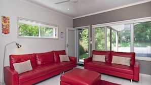 Una sala de estar moderna con dos sofás de cuero rojo, un pequeño reposapiés rojo y una lámpara de pie. Amplias ventanas ofrecen vistas a la vegetación exterior.