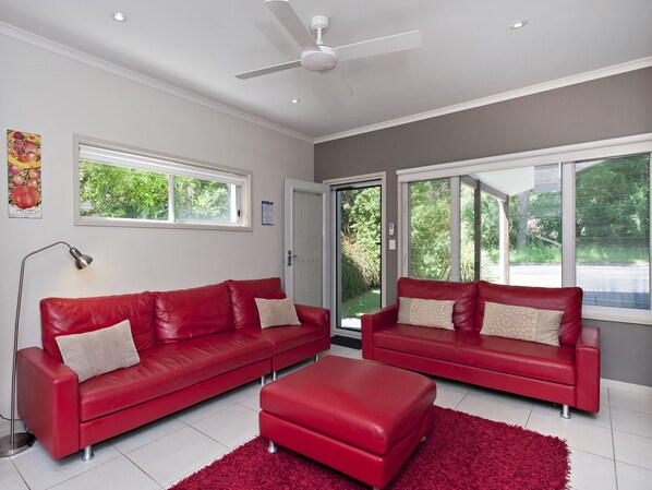 A modern living room with two red leather sofas, a matching ottoman, a ceiling fan, and large windows offering a view of greenery outside.